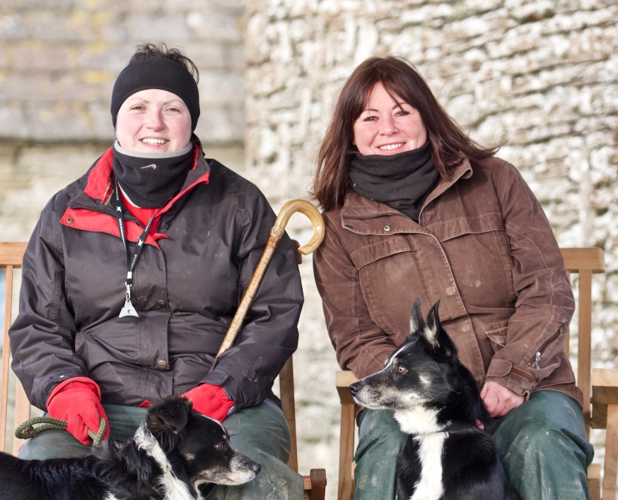 Image of two Women in Agriculture sitting side by side smiling. Sittting in front of them are two sheep dogs.